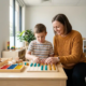 Ergotherapeutin kniet neben einem Kind beim praktischen Lernübung am Holztisch, bunte Lernmaterialien, helles Therapiezimmer.