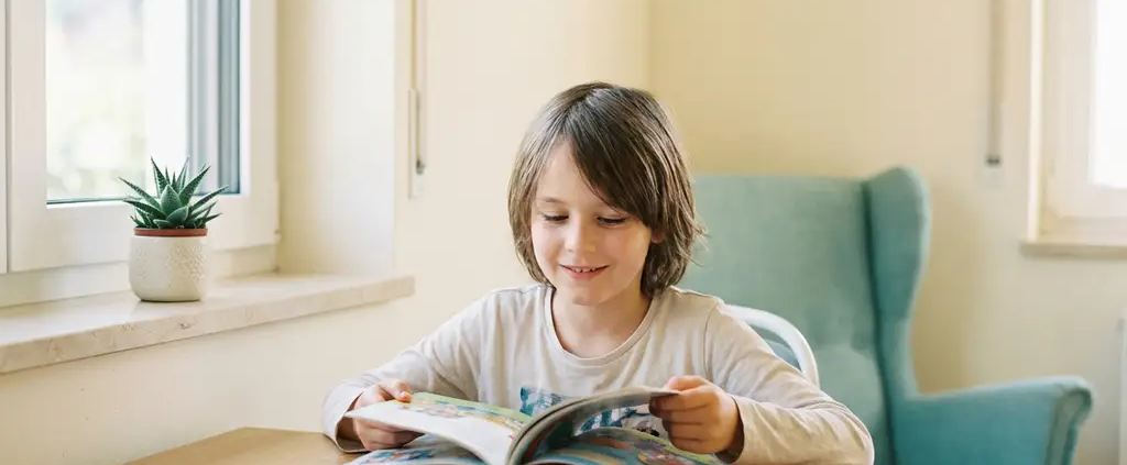 Kind liest konzentriert ein buntes Buch an einem hellen Holzschreibtisch mit natürlichem Fensterlicht und kleiner Pflanze.