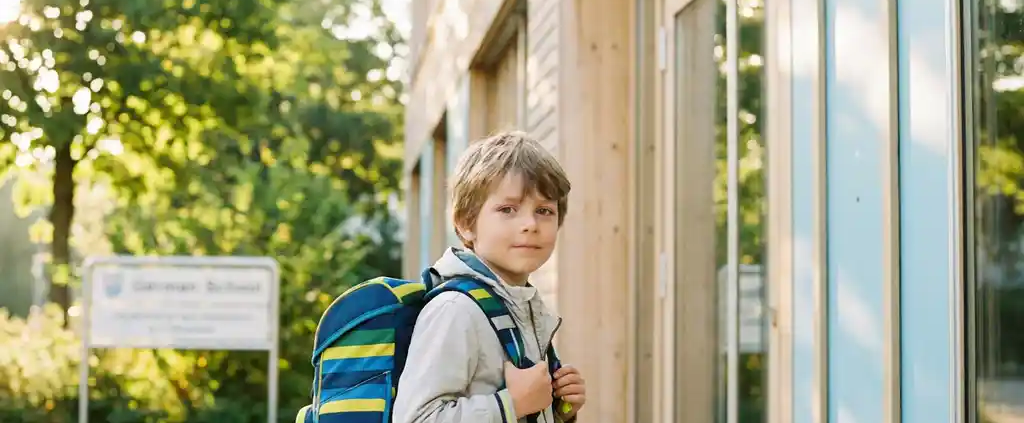 Kind mit Rucksack steht zögerlich vor einem modernen deutschen Schulgebäude im warmen Morgenlicht.