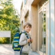 Kind mit Rucksack steht zögerlich vor einem modernen deutschen Schulgebäude im warmen Morgenlicht.