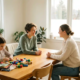 Therapeutin und Mutter im Gespräch an hellem Holztisch, Kind arbeitet mit bunten Materialien, skandinavisches Therapiezimmer mit Tageslicht.